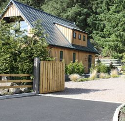 A wooden house with a metal roof behind a wooden gate and surrounded by trees and plants at Larchwood Lodge in Ellon