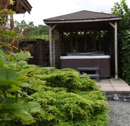 An outdoor patio area with a hot tub under a wooden gazebo surrounded by green bushes at Hillside View in Rhayader