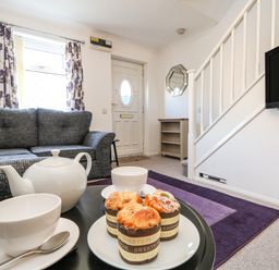 A living room with a gray sofa white teapot cups muffins a purple rug a wall-mounted tv and white stairs at Kingfisher Cottage in Weymouth