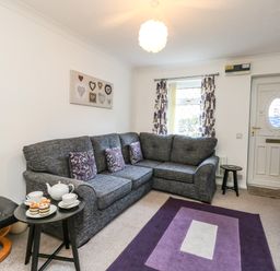 A living room with a gray sectional sofa and purple cushions a black side table with tea set and cupcakes a black footrest a purple rug a window with floral curtains framed hearts wall art and a white front door at Kingfisher Cottage in Weymouth
