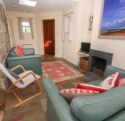 A living room with stone wall a wooden door two green sofas a rocking chair a fireplace and paintings at Rospletha Bungalow in Treen