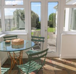 A dining area with a round glass table and green metal chairs near glass doors at Rospletha Bungalow in Treen