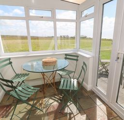 A sunroom with a glass round table and four green metal chairs overlooking a grassy field at Rospletha Bungalow in Treen