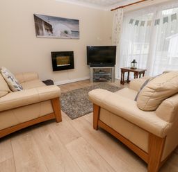 A living room with two beige sofas a wall-mounted fireplace a television on a stand a small wooden table and large window with curtains at 17 River Banks in Liskeard