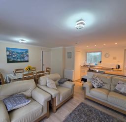 A living area with beige sofas and cushions a dining table with chairs and flowers and a kitchen with a window at 17 River Banks in Liskeard