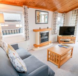 A living room with a grey sofa, wooden coffee table, television on wooden stand, fireplace, and windows with patterned curtains at Smugglers Shack in Polperro