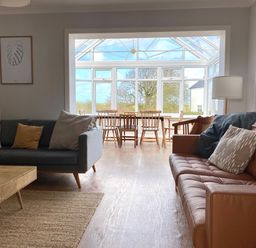 A living room with two sofas and a wooden table leading to a dining area with wooden chairs in a glass conservatory at Auverne in Poundstock near Widemouth Bay