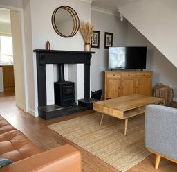 A living room with a wood stove fireplace a brown leather sofa a wooden coffee table a gray armchair and a wooden cabinet with a TV at Auverne Poundstock near Widemouth Bay