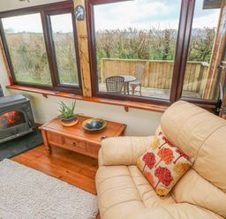 A living room with a leather sofa a wood burning stove a wooden table and large windows overlooking a wooden deck with chairs at Sarngwm in Narberth