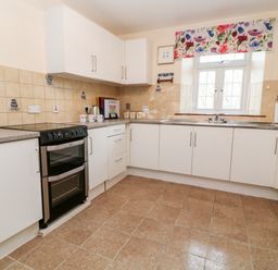 A kitchen with white cabinets, a black oven, a sink below a window with floral curtain, and beige tiled floor at The Vale in Ashwater