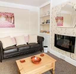 A living room with a brown leather sofa a wooden coffee table with bowls a wall-mounted electric fireplace and a built-in shelving unit at The Vale in Ashwater
