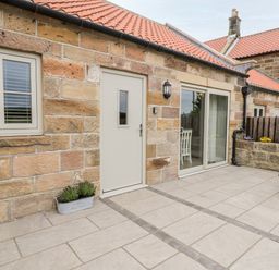 A stone cottage exterior with a beige door window and sliding glass door leading to a patio with a wooden picnic table and planter boxes at Shipswheel Cottage at Broadings Farm Whitby