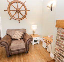 A brown armchair with a cushion beside a wooden side table with a lamp and nesting tables near an exposed brick fireplace at Shipswheel Cottage at Broadings Farm Whitby