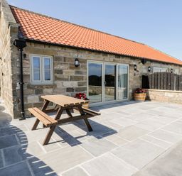 A stone building with a red tiled roof a wooden picnic table and flower pots on a paved patio at Stag Cottage at Broadings Farm Whitby