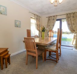 A dining room with a wooden table and six chairs with cushions a vase with flowers on the table curtains and glass doors leading outside at Rhiangwyn in Llanrhyddlad near Cemaes Bay