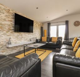A living room with black leather sofas and yellow cushions with a mounted TV on a stone wall at Box Tree Escapes in Kirkby Lonsdale