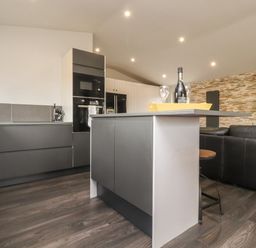 A kitchen with a dark grey island and cabinets a yellow kettle and bottle with glasses on the counter and a black sofa in a living area at Box Tree Escapes Kirkby Lonsdale