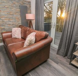 A living room with a brown leather sofa and two textured pillows next to glass doors with curtains and a small shelving unit at Box Tree Escapes Wennington near Kirkby Lonsdale