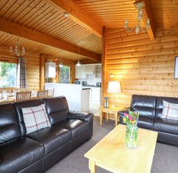 A living room with brown leather sofas and a wooden coffee table with flowers and a dining area with wooden chairs and a kitchen in the background at Valley View in Rhayader