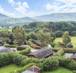 A cluster of small wooden cabins surrounded by trees and hedges with rolling hills and fields in the background at Mountain View in Rhayader