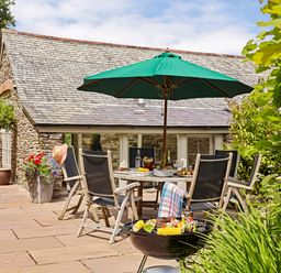 An outdoor patio area with a dining table and chairs under a green umbrella and a barbecue grill with food in front of a stone building at Garden House in Cornworthy