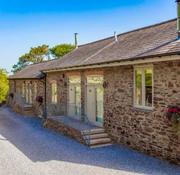 An exterior view of a stone building with multiple windows and doors along a gravel pathway with flower baskets attached to the wall at Haytor Cottage in Cornworthy