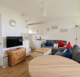 A living area with grey sofas with red and blue cushions a wooden coffee table and a round wooden dining table next to a kitchen area with white cabinets and a TV on a wooden stand at Pebbles in Cromer