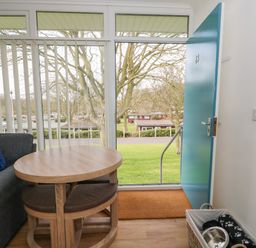 A living room area with a wooden table and cushioned stools next to a blue door with a view of trees and grass at Pebbles in Cromer