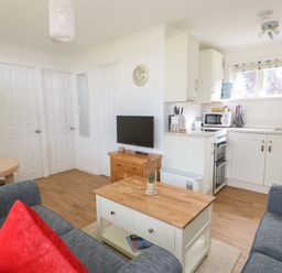A small living area with blue sofas red and blue cushions a wooden coffee table and kitchen with white cabinets at Pebbles in Cromer