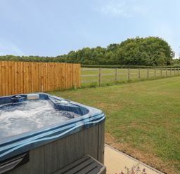 An outdoor hot tub next to a wooden fence and grassy yard with trees in the background at Buttercups at Midway in Kington