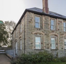 A stone building with multiple white a windows and a chimney with a paved pathway and metal railing at Stores & Supplies in Bodmin