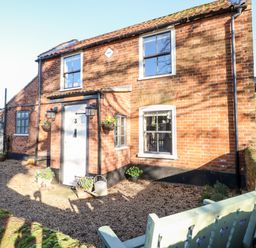 Exterior view of a brick cottage with white door and windows a gravel yard and a bench at Foundry Cottage in Reedham