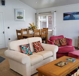 A living room with a beige sofa and colorful pillows a red armchair with matching ottoman a wooden coffee table and dining table with chairs at Lodge 11 in Tavistock
