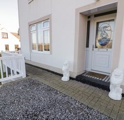 The front entrance of a house with a white door featuring a stained glass image of a ship two white lion statues on either side of the door a stone pathway and a white railing fence at Halcyon Moelfre near Benllech