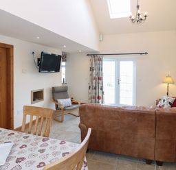 A living room with a brown leather sofa a wooden chair a dining table with a heart patterned tablecloth a wall-mounted television and double doors with curtains at Bryn Eiddon in Aberdaron