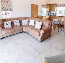An open living room and kitchen with a brown leather corner sofa with cushions a wooden dining table with chairs and kitchen cabinets at Bryn Eiddon in Aberdaron