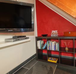 A mounted television above a white shelf with electronic devices next to a black cubed bookshelf with books and decorative items on a red wall at Y Bwthyn Newport in Newport Pembrokeshire