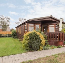 A wooden cabin with windows and a small porch surrounded by bushes and a lawn at 4 Country View Park in Graveney near Seasalter