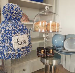 A tea set with a blue and white knitted tea cozy scones under a glass dome jam jars and stacked blue cups and saucers on a glass table at Valley View in Mevagissey
