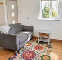 A small living area with a gray sofa, a side table with a wicker drawer, a colorful patterned rug, and a window showing trees at Valley View in Mevagissey