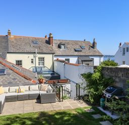 An outdoor patio with a sectional sofa and cushions next to a barbecue grill with houses and a sea view in the background at The Water House in Falmouth