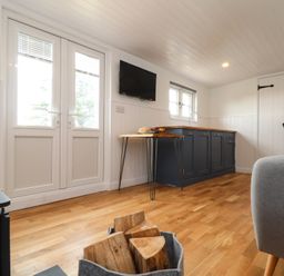 A living room with wood flooring a grey chair a basket of firewood a small table blue kitchen cabinets and French doors at Sebright in St Austell