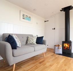 A room with a grey sofa with cushions a wood burning stove and a basket of firewood at Sebright in St Austell
