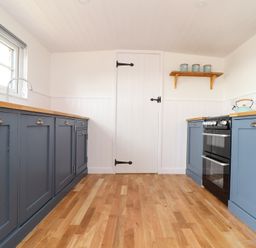 A kitchen with blue cabinets wooden countertops an oven a kettle a wall clock and a door at Sebright in St Austell