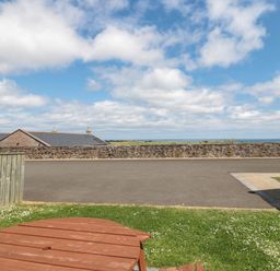 A grassy backyard with a wooden picnic table and a view of a stone wall and rooftops under a partly cloudy sky at Stable D'or in Embleton