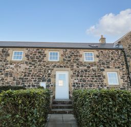 Front entrance of a stone cottage with a white door and windows surrounded by hedges at Low Croft in Embleton
