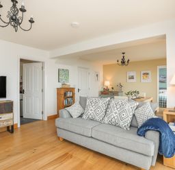 A living room with a grey sofa and patterned cushions a wooden TV stand with a TV an open door a wooden bookshelf a dining area with a chandelier and framed pictures at Red Ridge in Llandanwg near Harlech