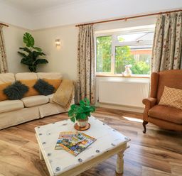 A living room with a beige sofa with cushions a brown armchair a white coffee table with magazines and a plant and floral curtains at Downs View in Norton Ferris