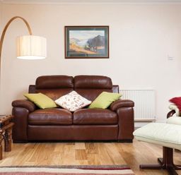 A living room with a brown leather sofa green and patterned cushions a wooden side table a curved floor lamp a framed landscape painting a cream armchair with a red cushion and a matching footstool at Nutwood in Lakeside