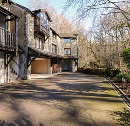 A driveway leading to garages of stone houses with trees and bushes nearby at Spring Cottage in Ambleside